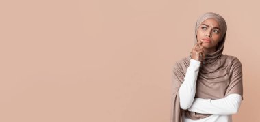 Portrait of doubtful afro muslim woman in headscarf touching chin, overthinking options and looking away over yellow background