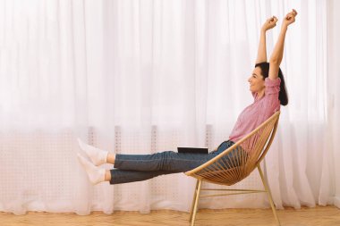 Weekends Finally. Young woman raising hands sitting in modern chair with a book on her lap, stretching body, copyspace clipart