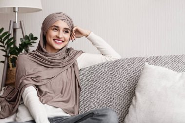 Güne gülümseyerek başla. Kafiyeli mutlu Arap kız portresi kanepede oturuyor ve mutlu bir yüz ifadesiyle boş yere bakıyor.