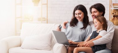 İnternet alışverişi. Üç kişilik neşeli bir aile internetten bilgisayar ve kredi kartıyla bir şeyler alıyor. Evdeki kanepede birlikte oturuyorlar.
