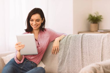 Teknoloji Konsepti. Kanepede oturan gülümseyen kız öğrenci, elinde tablet, video izliyor, arkadaşlarıyla sohbet ediyor.