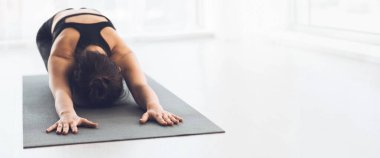 Bir kadın iyi aydınlatılmış bir stüdyoda yoga minderinde çocuk pozu veriyor, rahatlama ve farkındalık üzerine odaklanıyor, boşluğu kopyalıyor.