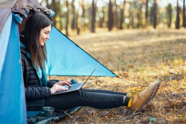 Çadırda tek başına oturan ve dizüstü bilgisayar kullanan neşeli kızın yan görüntüsü.