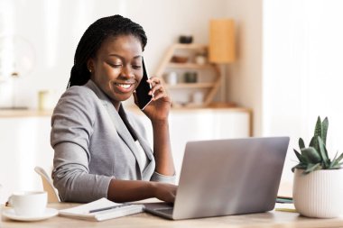 Afrikalı Amerikalı iş kadını cep telefonuyla konuşuyor ve modern ofiste dizüstü bilgisayar kullanıyor.