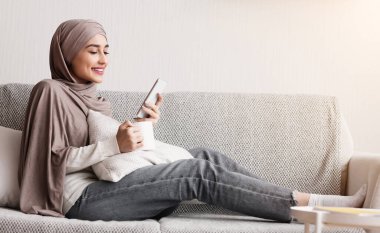 İslami Ev Kadınları Konsepti. Genç Müslüman kadın tesettürde, evde akıllı telefon ve kahveyle dinleniyor.