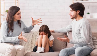 Aile sorunları kavramı. Ağlayan küçük bir kız evdeki kanepede tartışan ebeveynler arasında oturuyor, yüzü ellerine gömülüyor, korkmuş ve bunalımda.