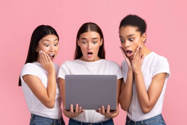 Üç Şaşırmış Milenyum Kızı dizüstü bilgisayara bakıyor ve Pembe Arkaplanda İnterneti inceliyor. Stüdyo Çekimi