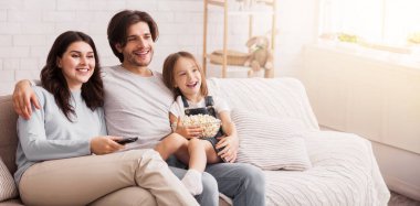 Aile eğlencesi. Neşeli ebeveynler evde kızlarıyla vakit geçirip televizyon izliyor ve patlamış mısır yiyorlar.