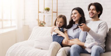 Baba, anne ve küçük kız evlerinde televizyon izliyor, rahat oturma odalarında koltuklarında oturuyor, baba kanalı değiştiriyor.
