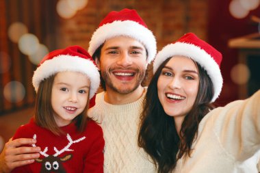 Noel Baba şapkalı üç kişilik neşeli bir aile Noel ışıklarında telefonla selfie çekiyor.