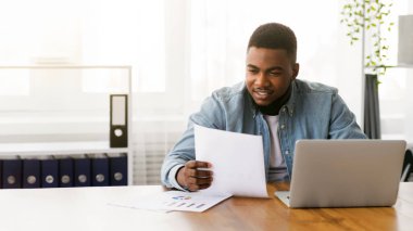 Gülümseyen Afrikalı Amerikalı iş adamı ofiste belgelerle çalışıyor, mali raporları kontrol ediyor..