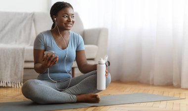Neşeli siyahi kadın, evde su ve akıllı telefonla idman yaptıktan sonra dinleniyor, gülümseyen Afrikalı kadın oturma odasında oturuyor, kulaklıkla müzik dinliyor, sağlıklı yaşam tarzının tadını çıkarıyor, boş alan arıyor.