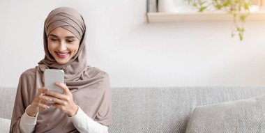 Portrait of millennial muslim girl in hijab using smartphone at home, messaging or browsing social networks, sitting on sofa in living room