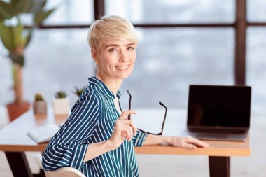 Ofis işi. Laptop 'ın başındaki neşeli olgun bayan modern ofiste oturan kamera pozuna gülümsüyor. Seçici Odaklanma