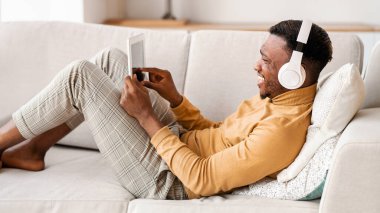 Müzik uygulaması. Kulaklıklı neşeli siyahi adam evdeki koltukta dinlenirken dijital tablet kullanıyor. Seçici Odaklanma