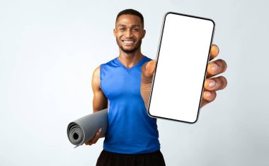 Fitness Konsepti. Elinde su şişesi ve yoga minderi tutan mutlu siyah adam, sarı duvarın üzerindeki kameraya gülümsüyor.