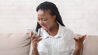 Öfkeli Afro Kadın, cep telefonuyla duygusal olarak bağırıyor, evde boş bir yerde, koltukta otururken biriyle telefonda tartışıyor.