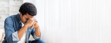 Afro-Amerikalı bir adam elleri bağlı ve başı eğik oturur, rahat bir oturma odasında derin duyguları yansıtır. Doğal ışık boşluğu doldurur, düşünceli atmosferi güçlendirir..