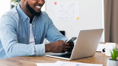 Modern teknolojiler iş başında. Gülümseyen afro girişimci akıllı telefon kullanıyor ve ofiste dizüstü bilgisayarla çalışıyor..