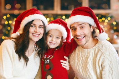 Kırmızı Noel Baba şapkalarıyla poz veren sevimli genç bir aile dekore edilmiş daire arka planında, kanepede kaynaşma, boş alan.