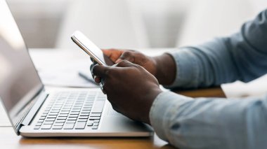 Tanımlanamayan afro iş adamı akıllı telefondan mesaj atıyor ve ofiste dizüstü bilgisayarla çalışıyor, kopyalama alanı ile yan görünüm
