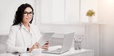 Meşgul Kadın. Kendine güveni olan Latin kız dizüstü bilgisayar kullanıyor, elinde tablet tutuyor, kameraya bakıyor, kopyalama alanına bakıyor.