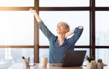Dizüstü bilgisayarda kollarını geren mutlu iş kadını iş yerinde kapalı bir yerde oturmaktan memnun. Seçici Odaklanma