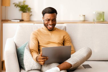 Neşeli Afrikalı Amerikalı adam dizüstü bilgisayar kullanıyor ve kulaklık takıp evdeki koltukta otururken video görüşmesi yapıyor. Boş Alan, Seçici Odaklanma