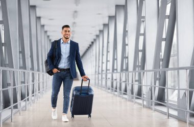 Havaalanı terminalinde bavulla yürüyen genç bir orta doğunun portresi, biniş kapısına giderken bavul taşıyan gülümseyen Arap adam, hava yolculuklarının ve iş seyahatlerinin tadını çıkarıyor, kopyalama alanı.