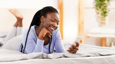 Pozitif siyah kız akıllı telefon kullanıyor. Sabahları yatakta uzanıyor. Seçici Odaklanma