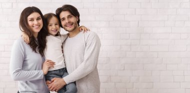 Caring Family. Portrait Of Loving Parents With Their Cute Little Daughter On Hands Posing Over White Brick Wall Background Indoors, Copy Space
