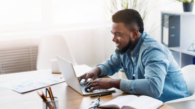 Modern iletişim. Gülümseyen Afro-Amerikan çalışanı müşteriye e-posta yazıyor, ofiste dizüstü bilgisayarla çalışıyor.