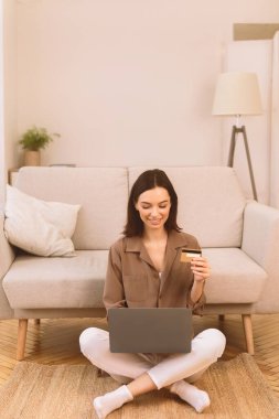 Çevrimiçi Mağaza. Mutlu genç bir kadın internet üzerinden alışveriş yapıyor. Laptop ve kredi kartı kullanıyor. Yerde oturuyor.
