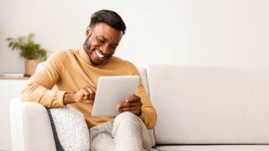 Evdeki koltukta oturmuş gülümseyen dijital tablet kullanan neşeli siyahi adam. Güzel Mobil Uygulama. Seçici Odaklanma