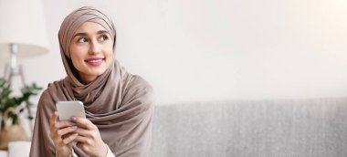 Kafiyeli, akıllı telefonlu düşünceli Arap bir kızın portresi. Evdeki koltukta oturuyor ve başka tarafa bakıyor.