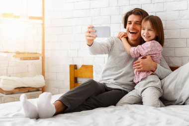 Mutlu anaokulu kızı ve babası akıllı telefondan selfie çekiyor, sabahları yatakta uzanıyor, boş yer var.