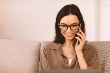 Gözlüklü genç bir kadın cep telefonuyla konuşuyor evdeki rahat koltukta oturuyor, dizüstü bilgisayara bakıyor, kopyalama alanına bakıyor.
