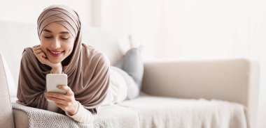 tesettürlü çekici Müslüman kadın evde rahat bir kanepede uzanırken akıllı telefon kullanıyor, sosyal ağlara mesaj atıyor ya da göz gezdiriyor, kapatıyor