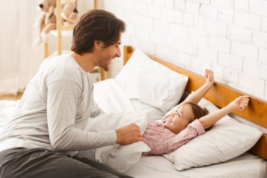 Anaokulu kızı uyandıktan sonra yatakta geriniyor. Genç babasına gülümsüyor.