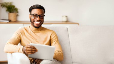 Hafta sonu. Gözlüklü Afro-Amerikan adam dijital tablet tutuyor ve evdeki koltukta oturan kameraya gülümsüyor. Seçici Odaklanma