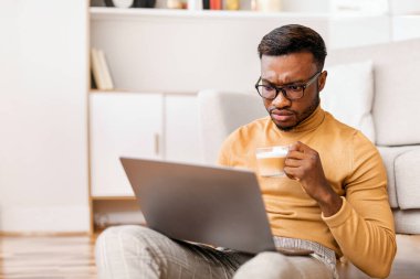 Ciddi bir Afro iş adamı dizüstü bilgisayar okuyarak iş haberleri okuyor. Sabah kahvesini evde oturarak içiyor.