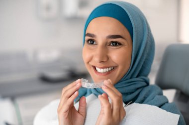 Ortodonti Konsepti. Tesettürlü Gülümseyen Müslüman Kadın Diş Hekimliği veya Görünmez Diş tellerini Klinikte Dişçi Sandalyesinde Otururken, İslami Kadın Diş Düzeltme İçin Temiz Diş Hattını Kullanıyor