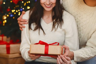 Noel Baba şapkalı neşeli adam, gülümseyen kadınını Noel hediyesiyle selamlıyor, Noel arifesini evde birlikte geçiriyor.