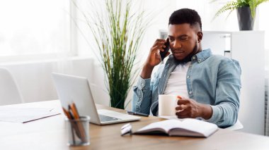 Çoklu görev konsepti. Milenyum Afro-Amerikan çalışanı cep telefonuyla konuşuyor, dizüstü bilgisayarla çalışıyor ve işyerinde kahve içiyor.