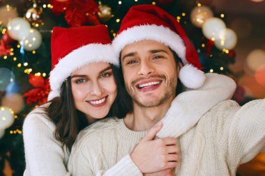 Noel arifesinde selfie çeken, Noel ağacının üzerine Noel Baba şapkası takan tatlı evli çift.