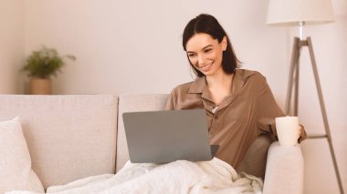 İyi sabahlar. Gülümseyen öğrenci kanepede oturmuş kahvesini yudumluyor ve dizüstü bilgisayar kullanıyor, video seyrediyor.