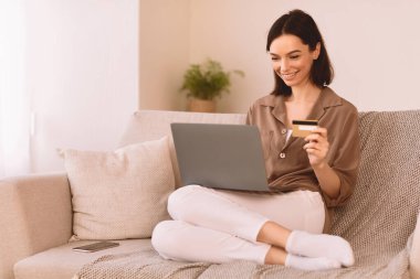 Çevrimiçi Mağaza. Mutlu kız internetten alışveriş yapıyor. Laptop ve kredi kartı kullanıyor. Kanepede oturuyor.