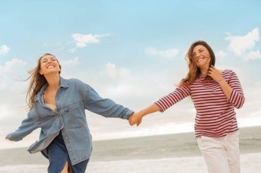 İki kız arkadaş mutlu bir şekilde kumsalda el ele yürüyor, gülümsüyor ve anın tadını çıkarıyorlar. Güneş arka planda batıyor, canlı ve sıcak bir atmosfer yaratıyor..