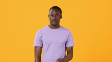 Genç adam parlak sarı bir arkaplan karşısında duruyor, basit bir mor tişört giyerken, güvenli bir şekilde el kol hareketi yapıyor. İfadesi ilgi çekici ve odaklı..