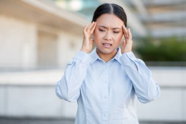 Genç bir Asyalı kadın şehir ortamının dışında duruyor, başını bir rahatsızlık bakışı ile tutuyor, muhtemelen bir baş ağrısı veya stresle uğraşıyor, profesyonel bir kıyafet giyerken..
